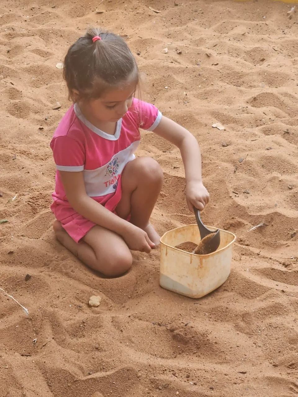 Menina brincando com uma pazinha e um balde na areia do parque da escola
