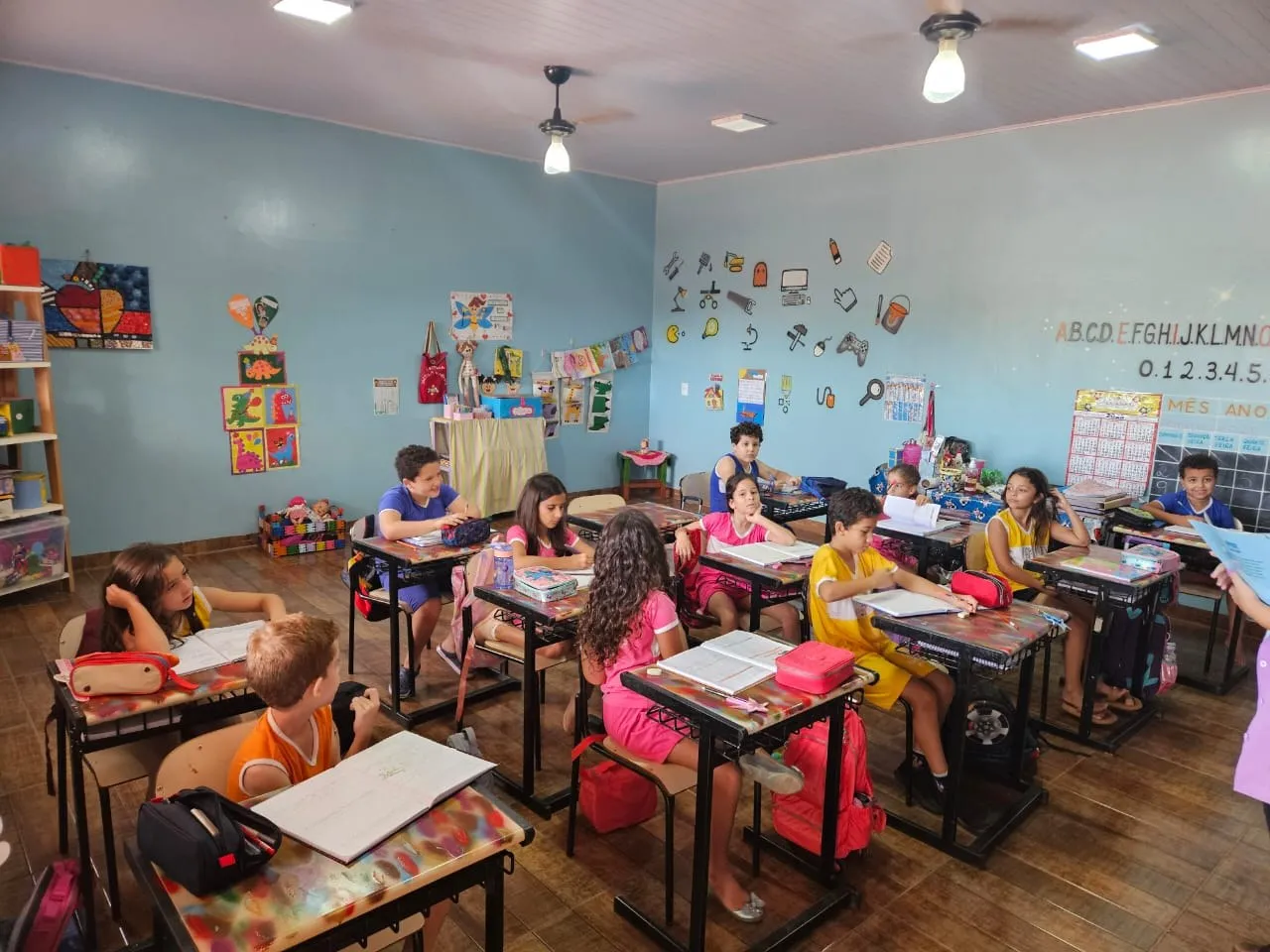 Alunos lendo em suas carteiras, em sala com alfabeto e numerais na parede