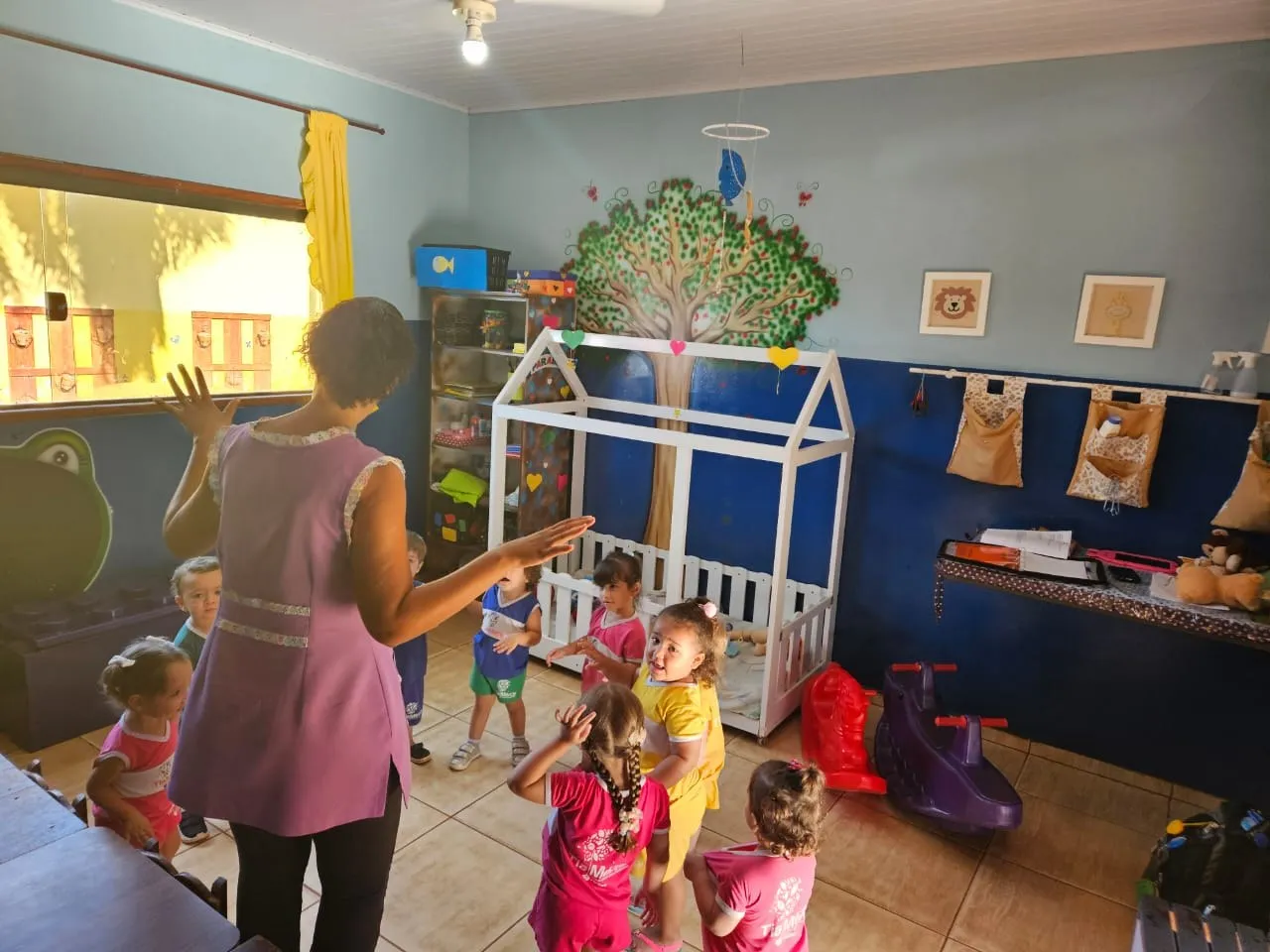 Professora cantando com as crianças pequenas ao lado de uma caminha de brinquedo