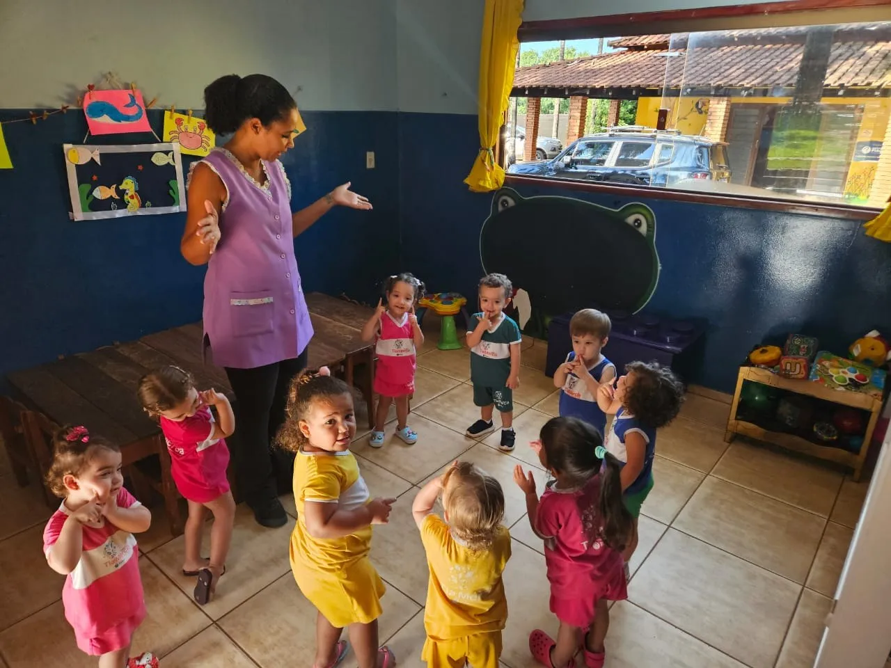 Crianças pequenas de uniforme amarelo e rosa em roda com a professora
