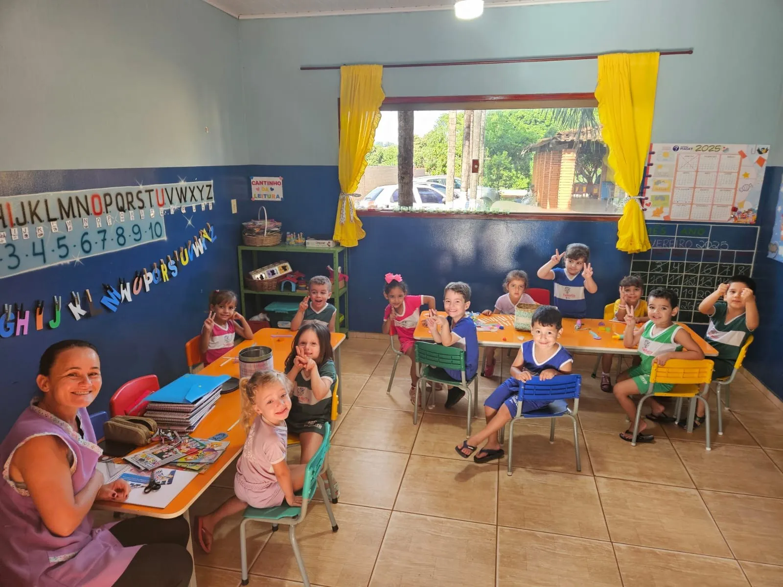 Crianças sentadas às mesinhas coloridas, com a professora sorrindo em primeiro plano