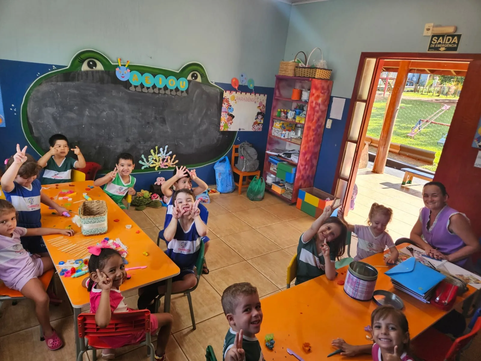Turma sorrindo para a foto nas mesas laranja, com a lousa em formato de sapo ao fundo