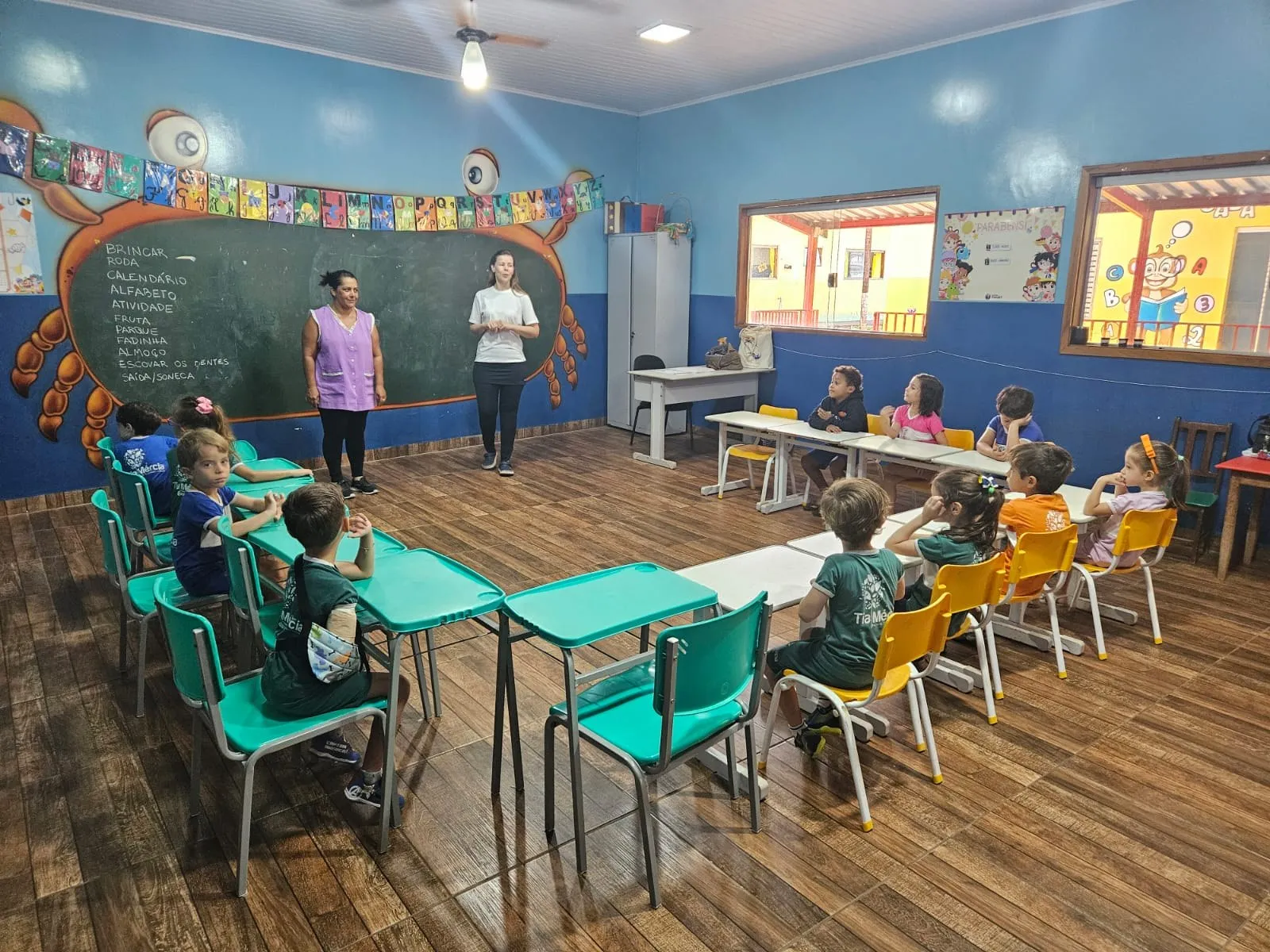 Duas professoras com a turma sentada em carteiras verde-água e amarelas