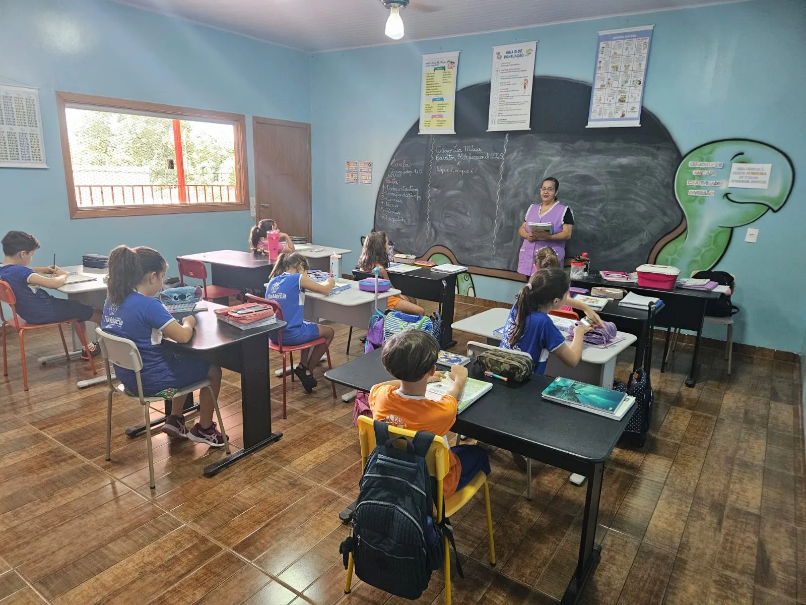 Alunos atentos à professora, sentados em carteiras escuras