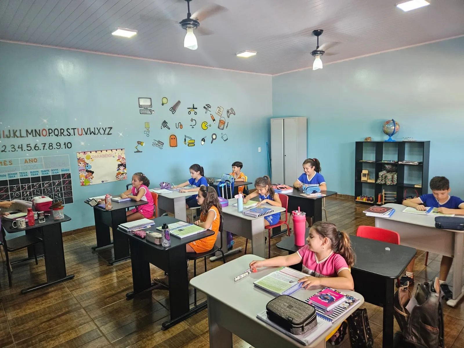 Alunas fazendo atividade, em sala com estante de livros e globo terrestre